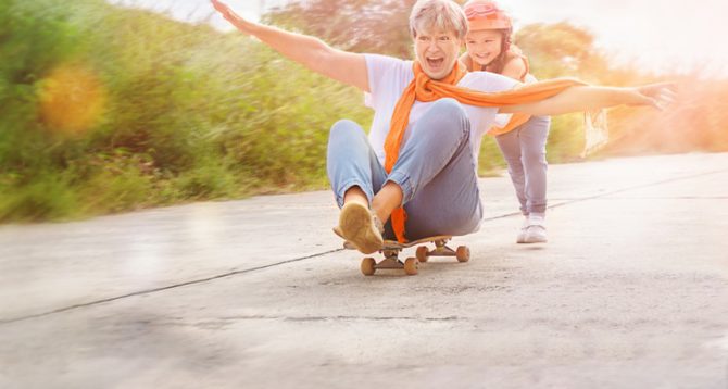 An older woman with gray hair, wearing an orange scarf and light blue jeans, sits joyfully on a skateboard, her arms spread wide. A young child with a helmet and a big smile stands behind her, pushing the skateboard. The scene takes place outdoors on a sunny day, with a blurred green background and a warm, happy atmosphere.