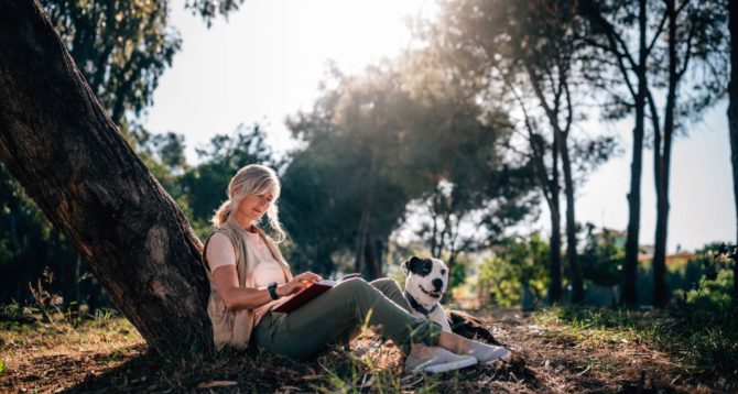 Fashionable senior woman with pet dog sitting under tree in forest park and reading book