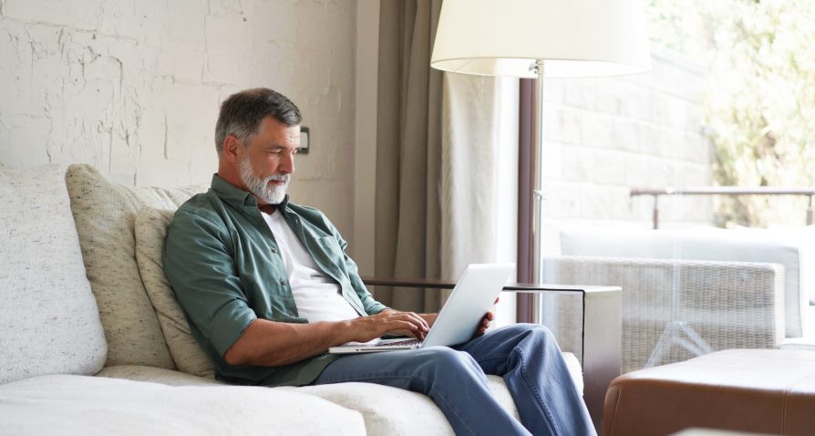 Senior man with gray hair and beard using a laptop while sitting comfortably on a sofa in a bright living room, researching Medicare plans. He is casually dressed in a green shirt and blue jeans, with a calm and focused expression. This image represents a relaxed and informed approach to choosing Medicare health plans.